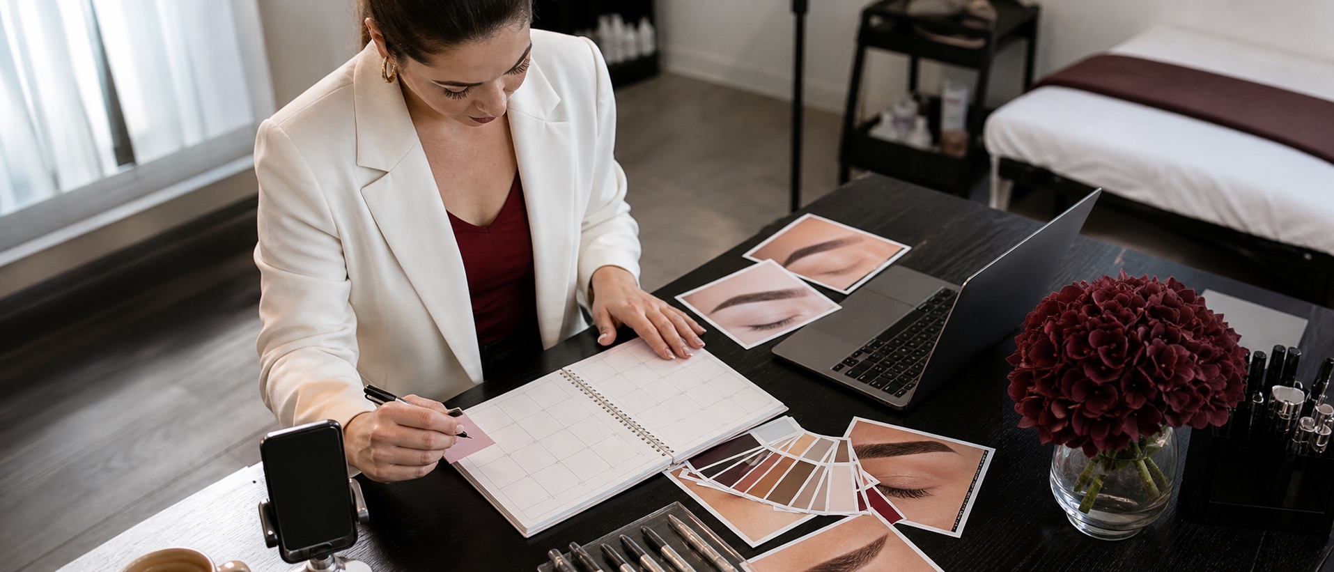 PMU artist planning a monthly content calendar with brow sketches, laptop and neutral studio stationery