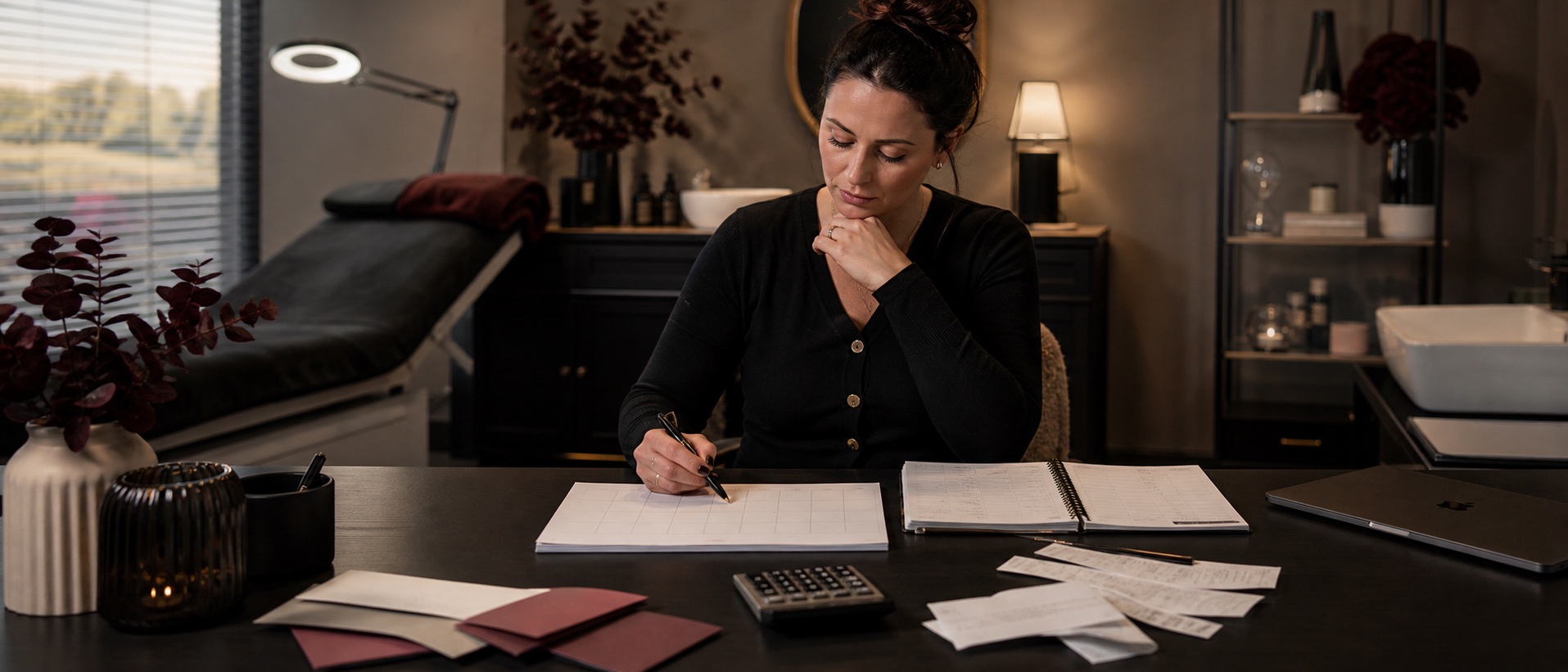 PMU studio owner reviewing a calendar, calculator and receipts at a desk beside a treatment room